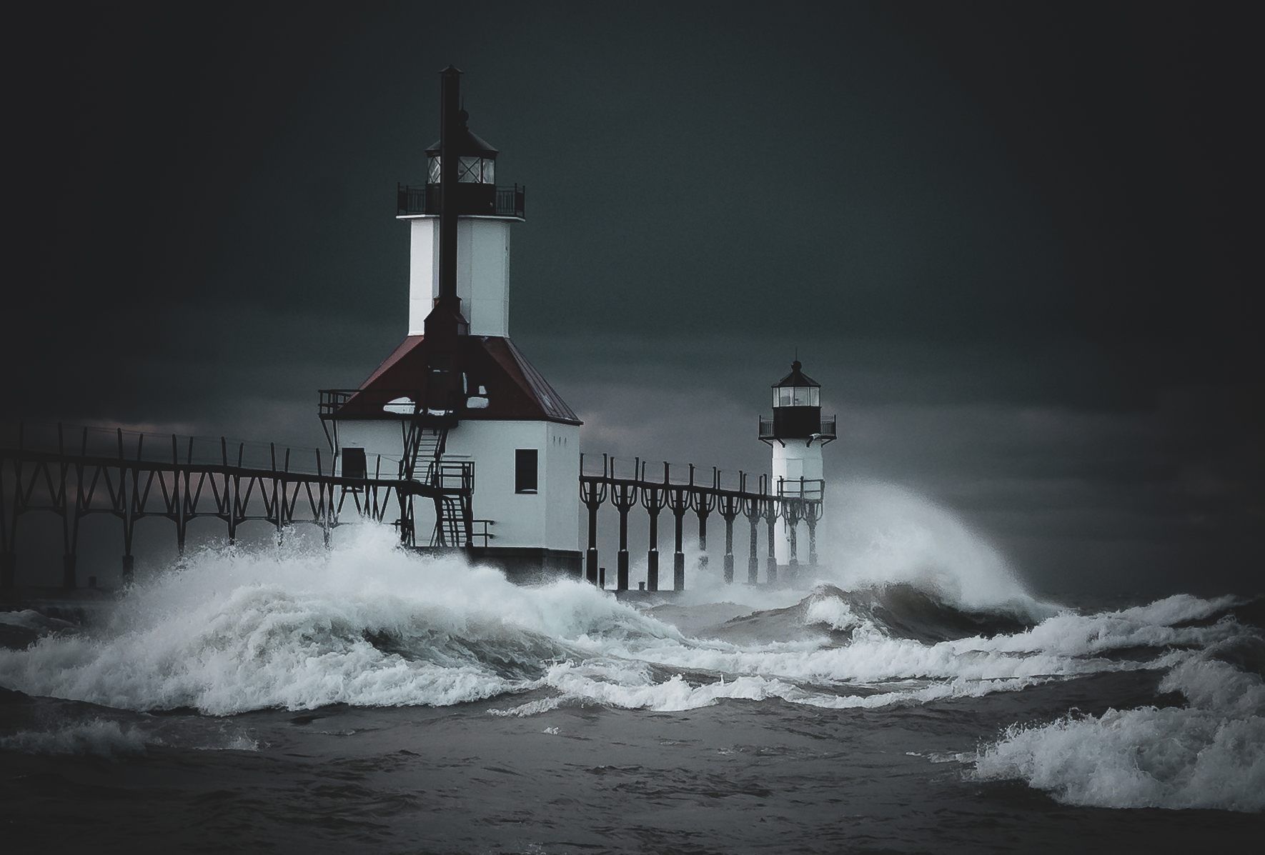 Two lighthouses on a storm-battered pier, large waves crashing around them under a dark sky