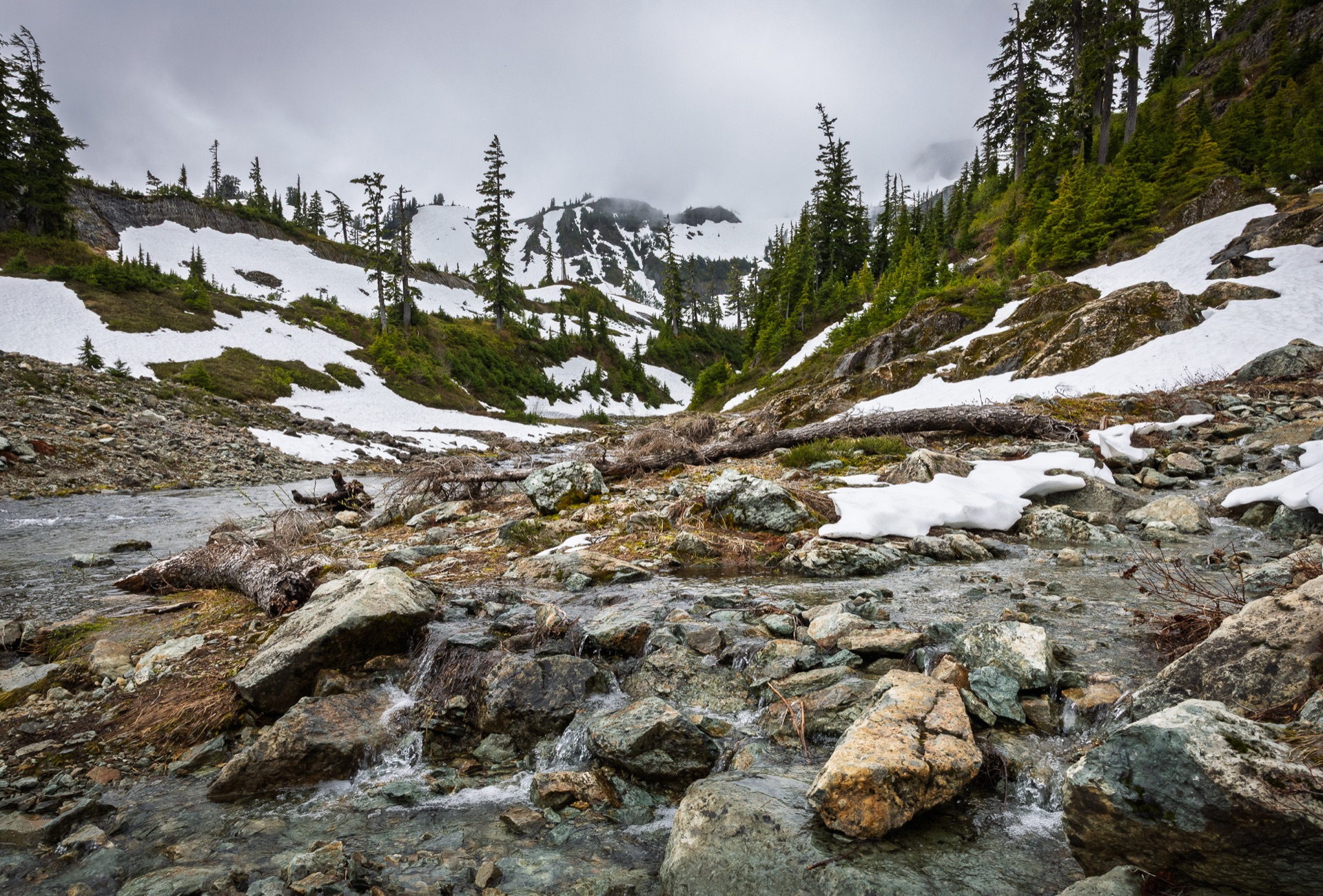 A stream running over rock through patches of snow below an alpine ridge